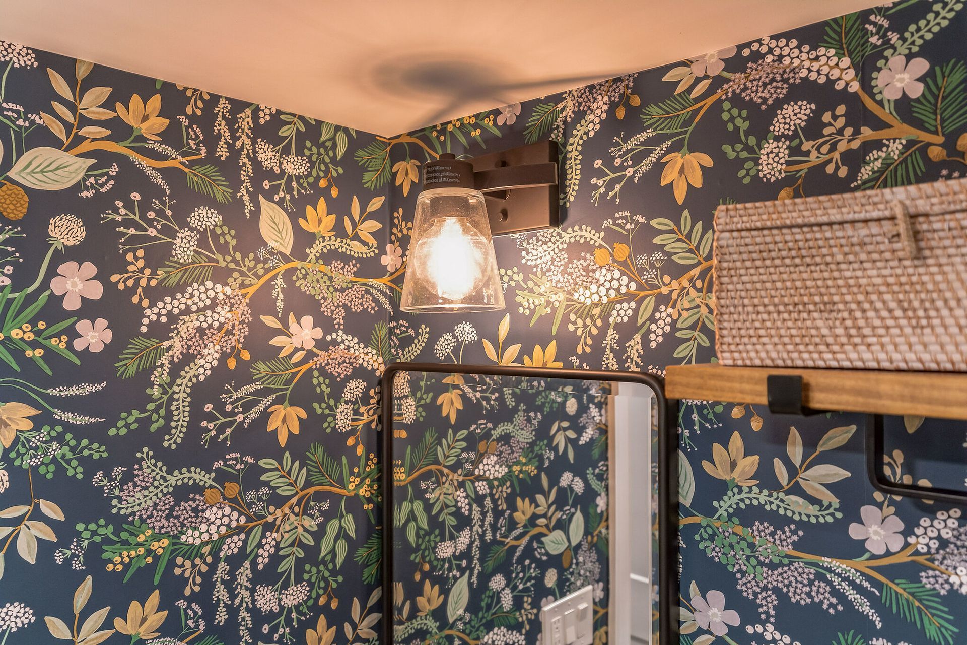 close-up of a clear glass sconce light on floral wallpaper with mirror and wood shelf partially visible
