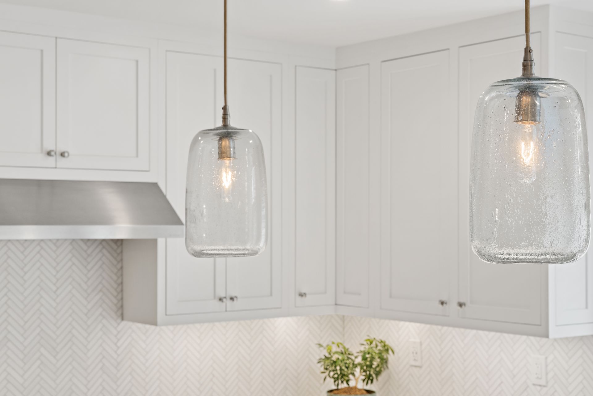 Glass pendant lights over white kitchen cabinets with herringbone tile backsplash and stainless steel range hood