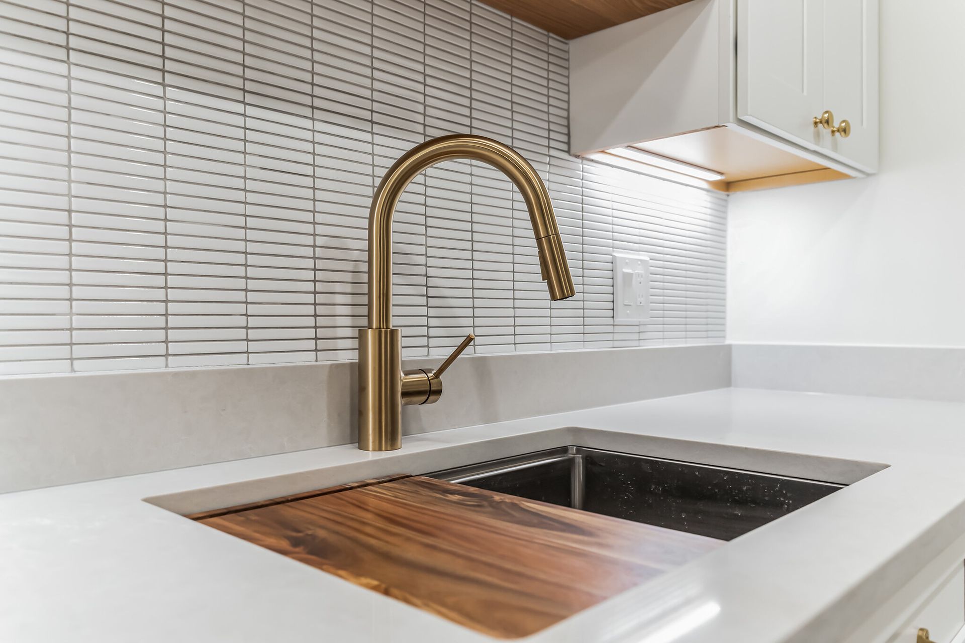 gold kitchen faucet close-up with cutting board sink, white quartz countertop, vertical tile backsplash, under cabinet lights
