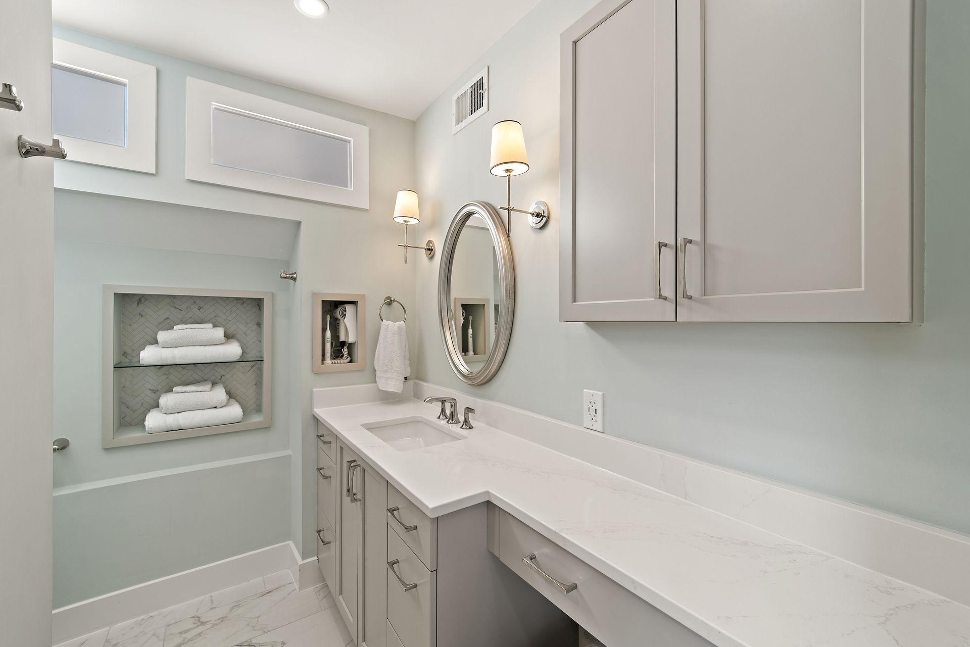 Gray bathroom vanity with round mirror, built-in shelving niches, and white quartz countertop