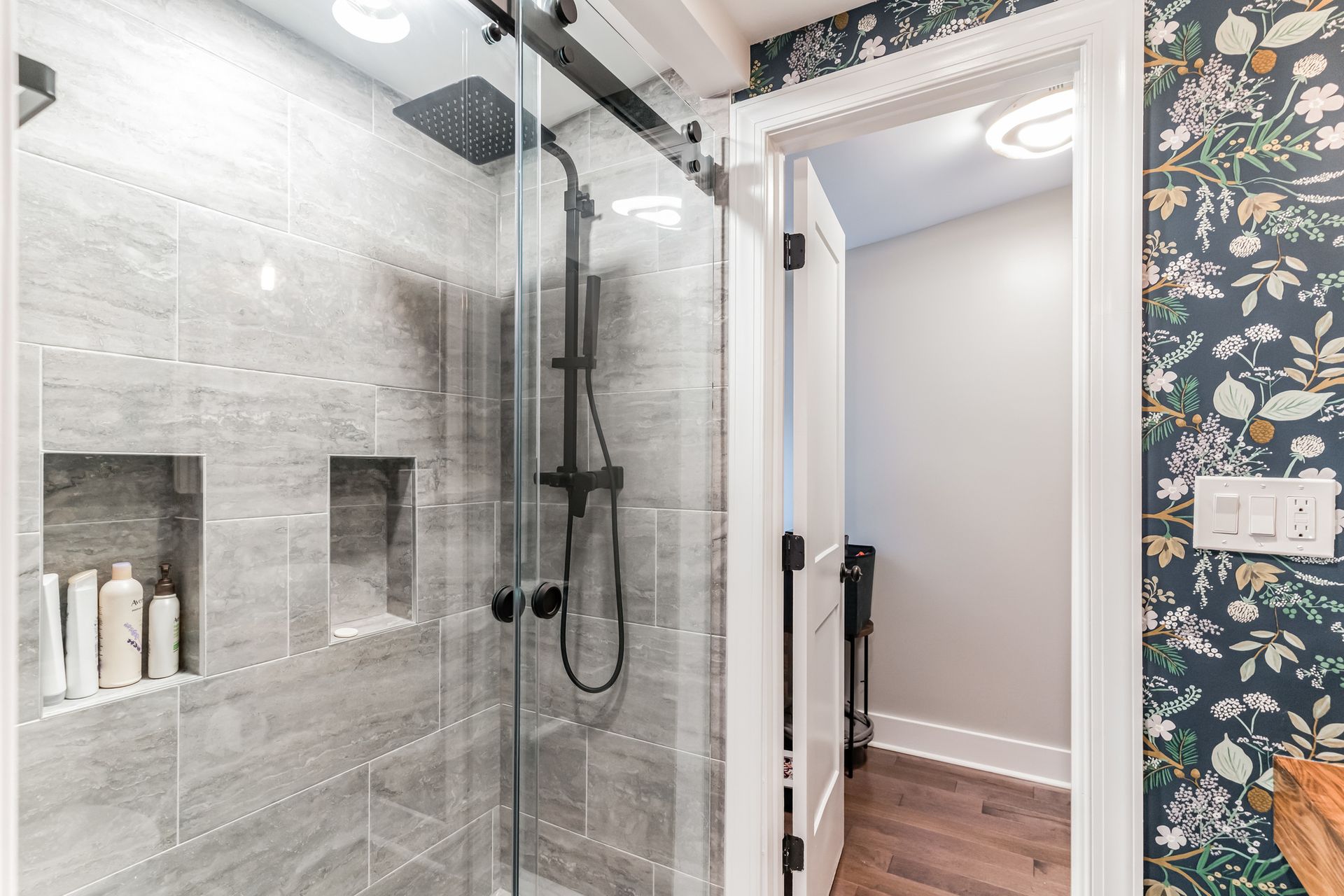 gray tile shower with black fixtures and sliding glass door next to floral wallpapered wall and white door