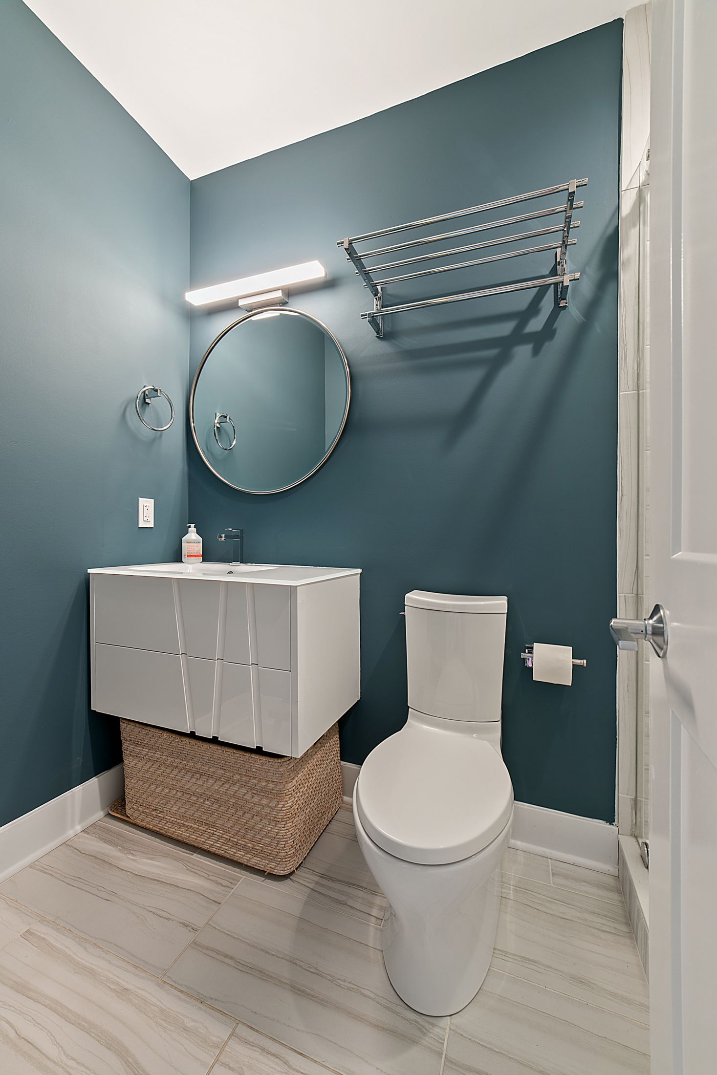 Modern hall bathroom remodel with blue wall, round mirror, white vanity, and marble-look tile floor in updated DC home