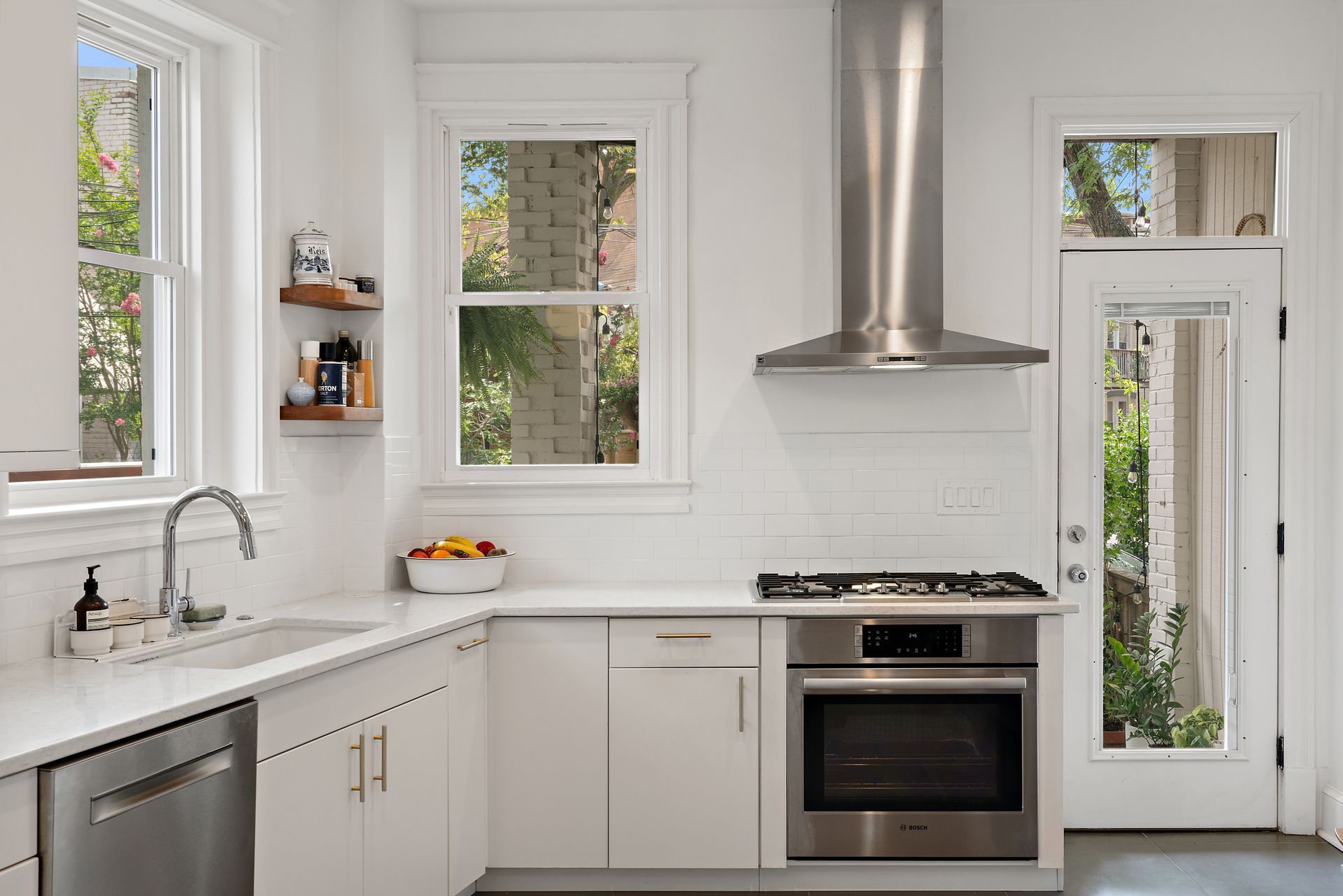 Bright white kitchen with corner sink under windows, open wood shelving, gas range, and stainless hood in Washington DC remodel