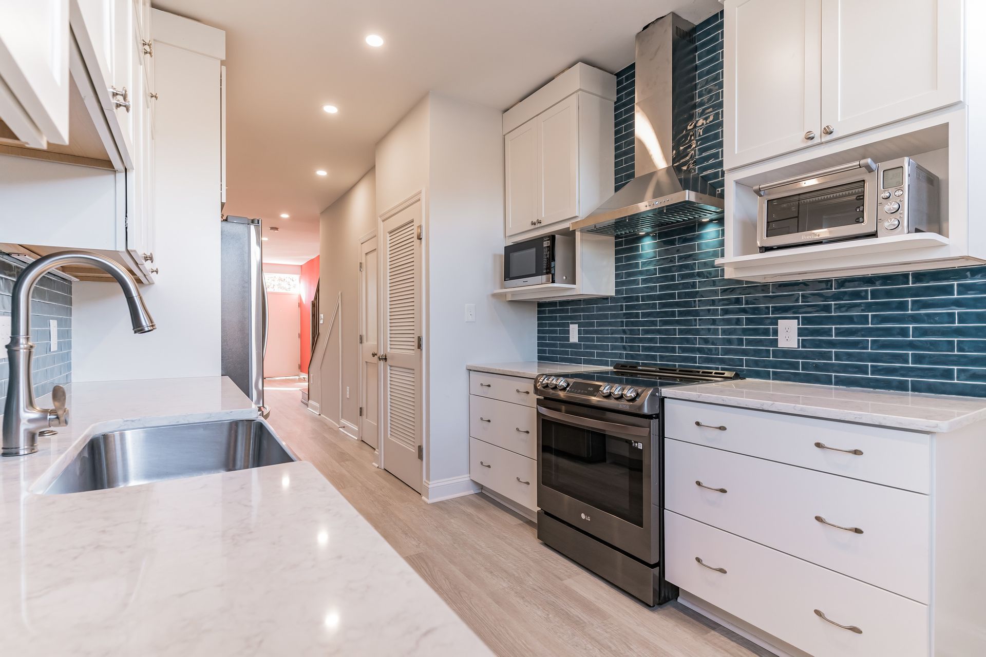 Modern galley kitchen with white cabinets, blue subway tile backsplash, stainless steel range hood, and light quartz counters