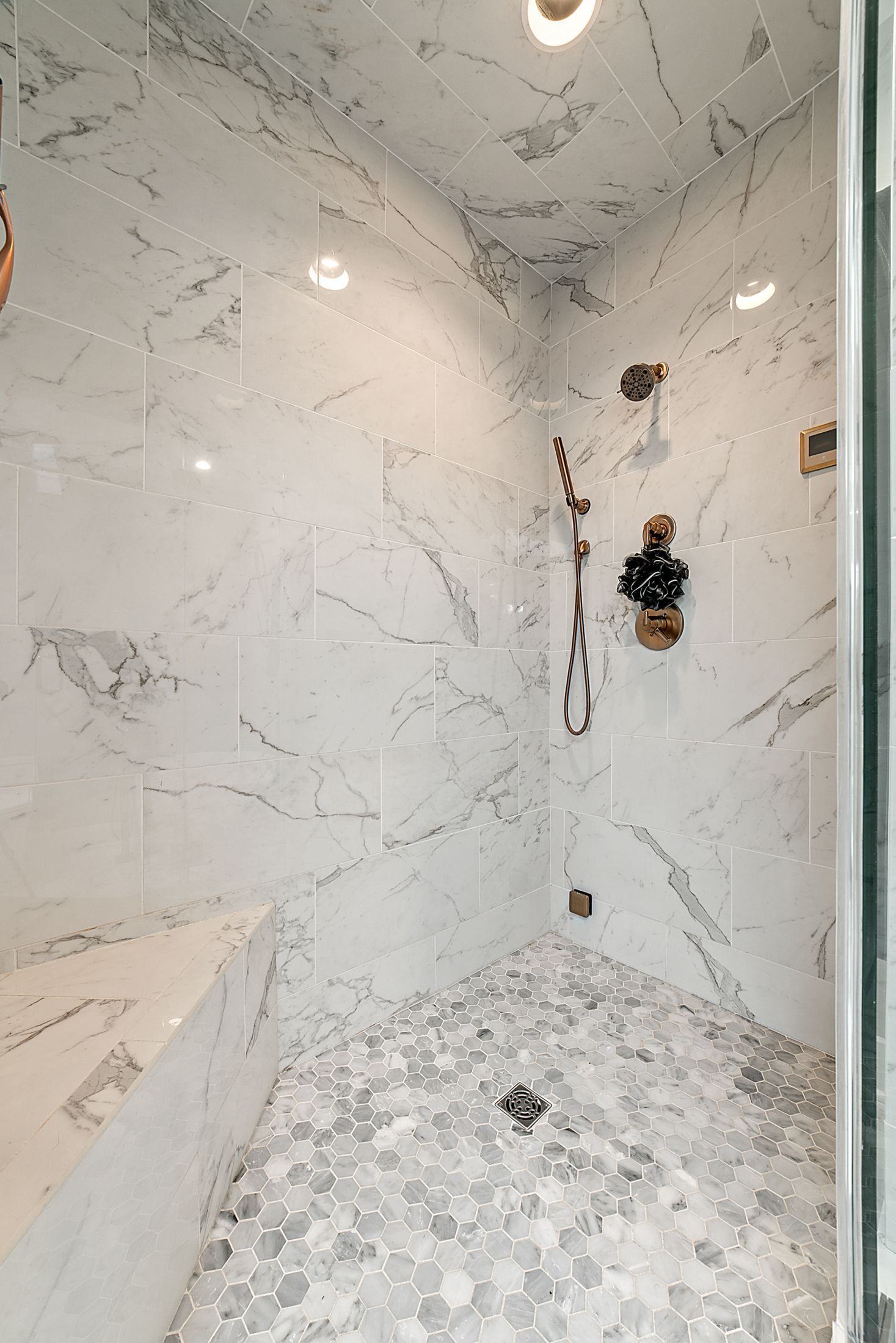 Marble walk-in shower with bench, white and gray hex tile floor, brass fixtures, and recessed lighting
