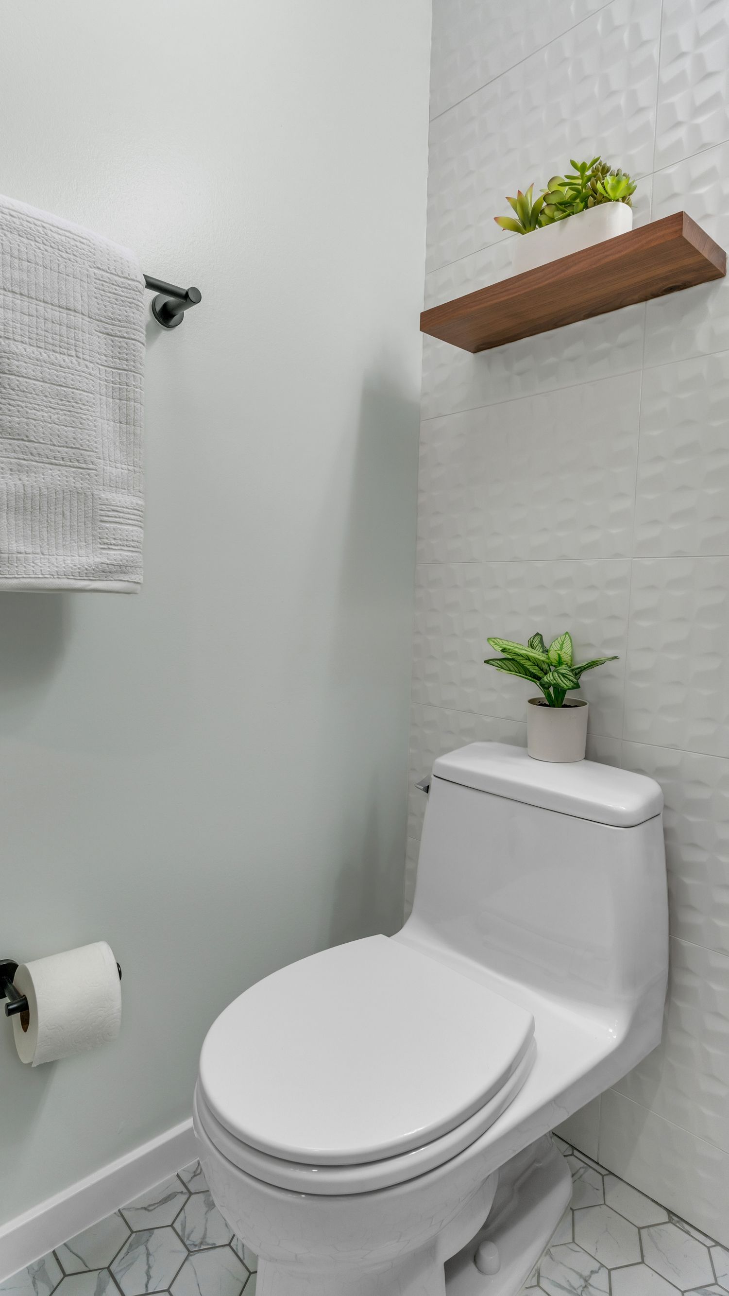 modern bathroom remodel with white toilet, floating wood shelf, textured wall tile, and hex marble floor tiles