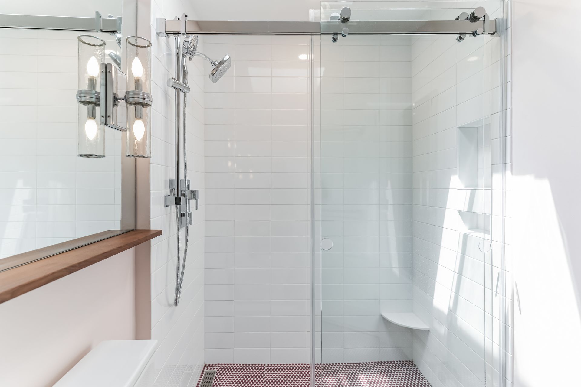 modern white tile shower with glass door, bench, handheld showerhead, and double sconce light