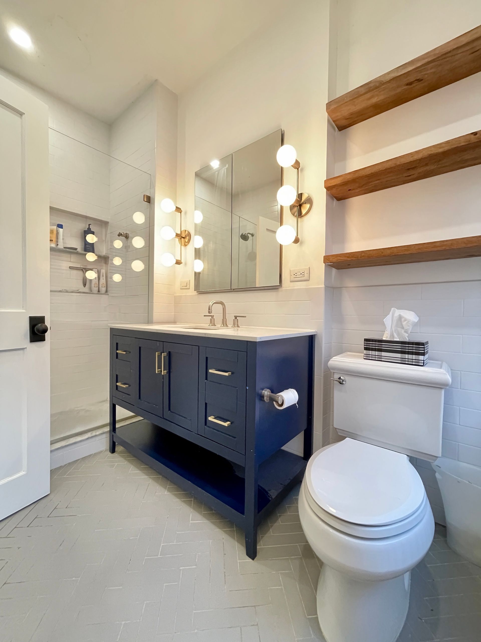 modern bathroom remodel with navy vanity brass lighting white subway tile and open wood shelving