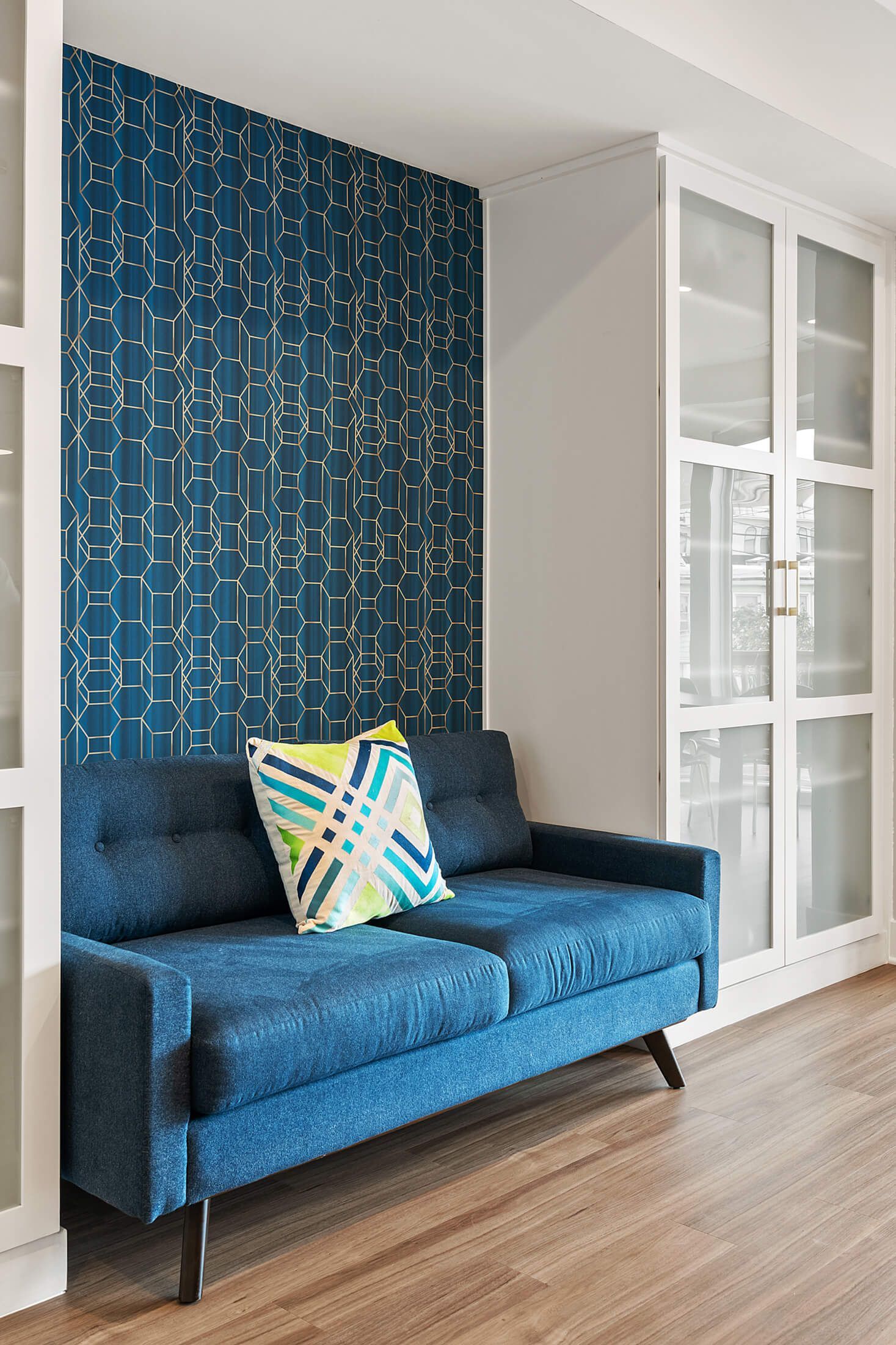 Kitchen sitting area with modern blue accent wall, glass cabinet doors, and mid-century style blue sofa with colorful pillow