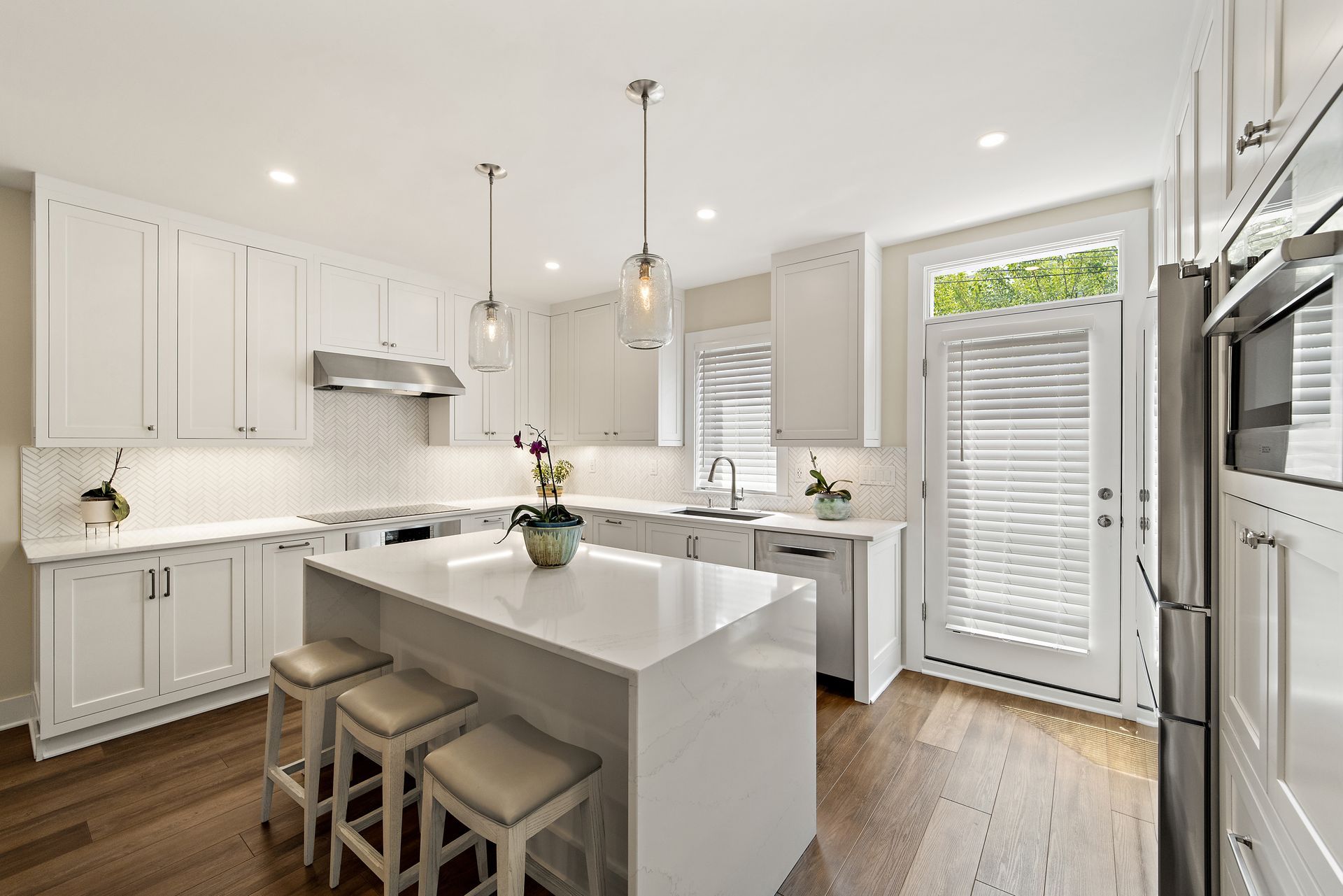 Transitional white kitchen featuring herringbone backsplash, waterfall island, glass pendant lighting, and natural wood flooring