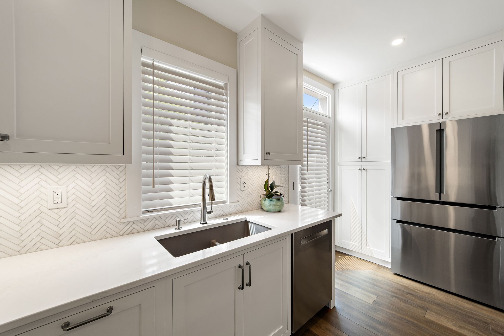 Bright white kitchen with herringbone backsplash, stainless steel refrigerator, and under-mount sink with brushed nickel faucet