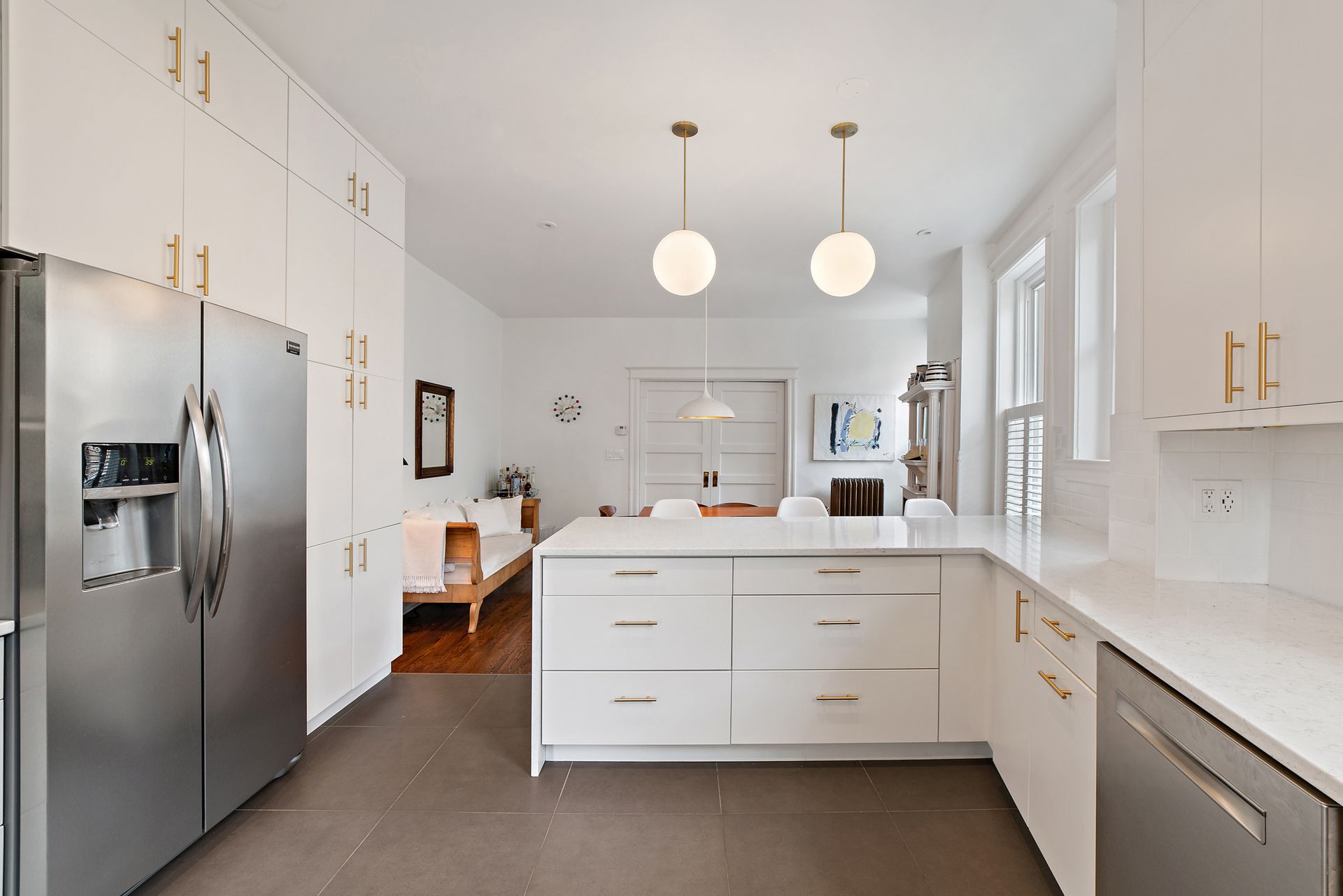 Bright white kitchen with quartz island, stainless steel appliances, gold hardware, globe pendant lights, and a view of an open dining and sitting area with eclectic décor.