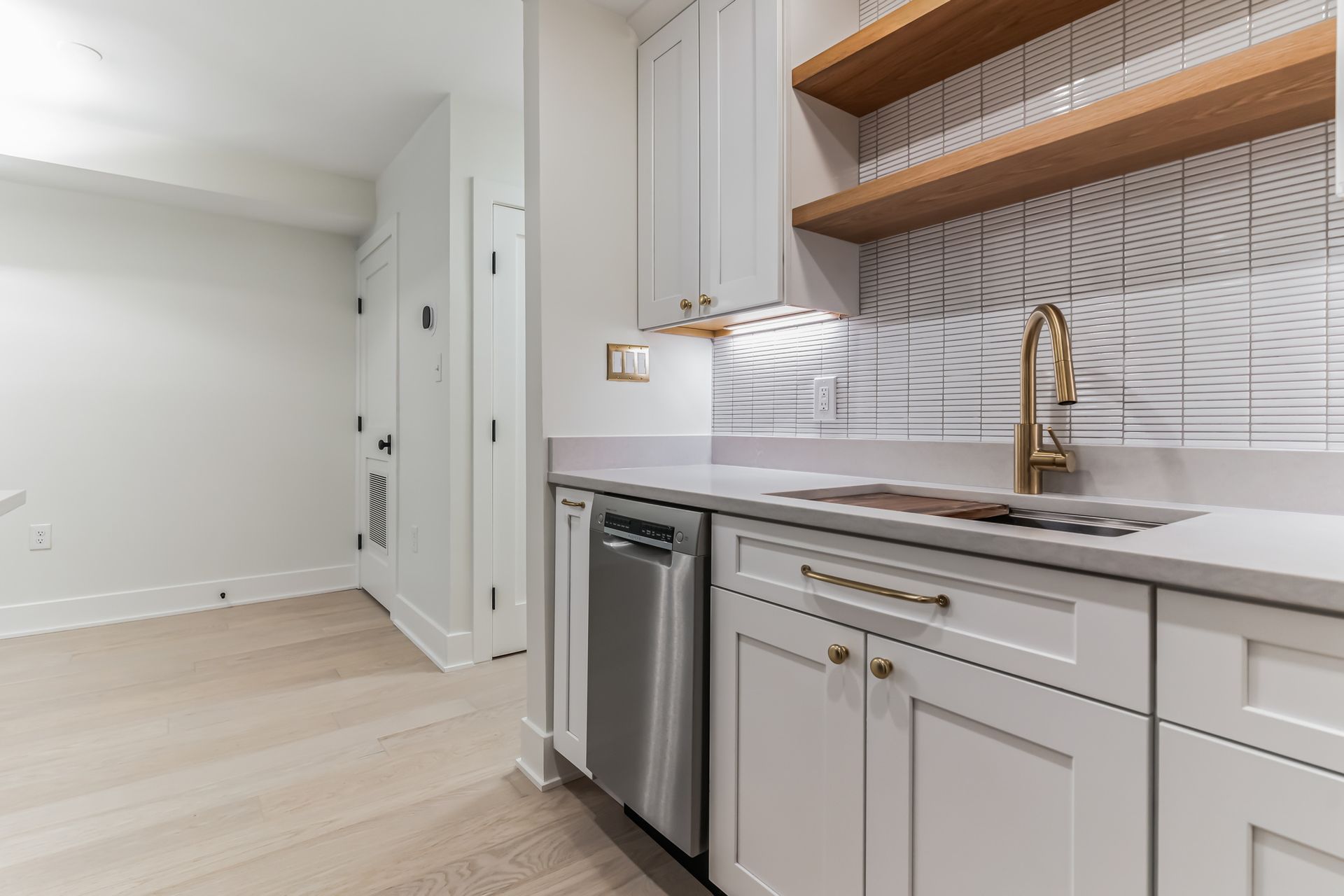 modern white kitchen remodel with gold faucet, open wood shelves, tile backsplash, under-cabinet lighting, and light wood floors