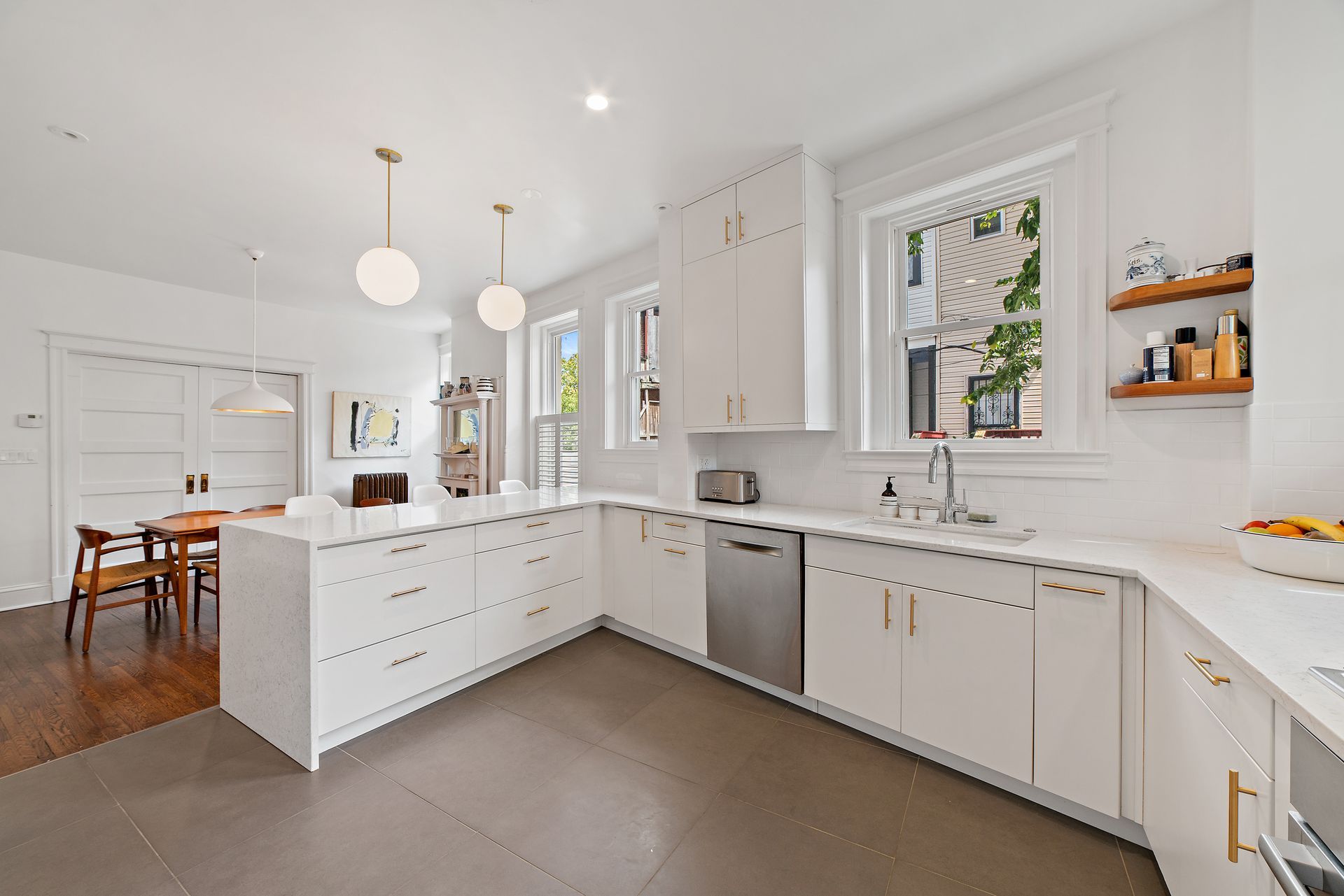 Spacious white kitchen with modern pendant lights, brass hardware, and wood and tile flooring, featuring a large island and open shelving.