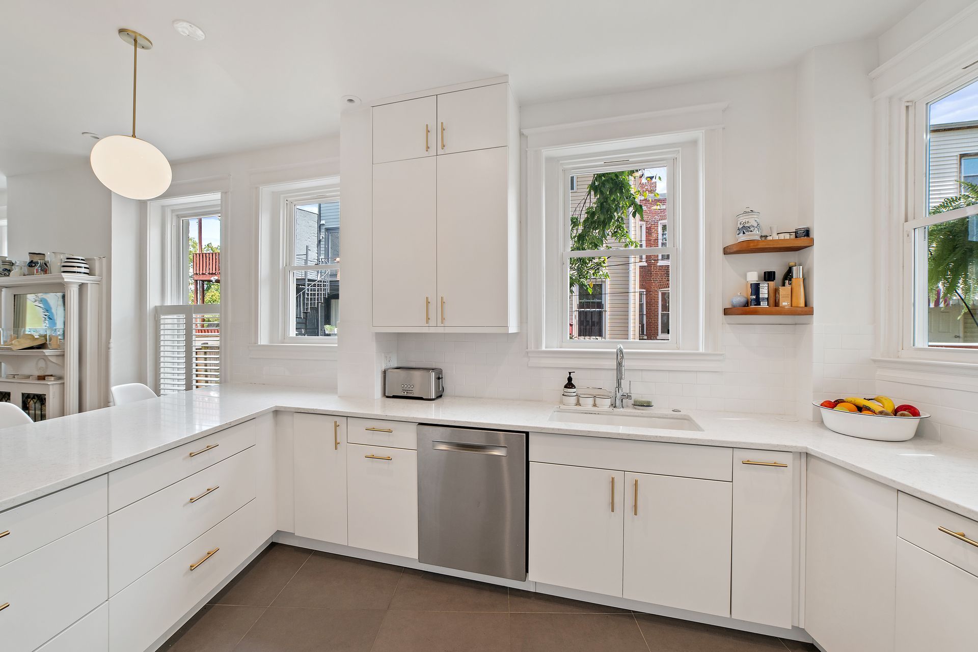 Bright U-shaped white kitchen with brass hardware, large windows, open wood shelving, and modern pendant lighting.