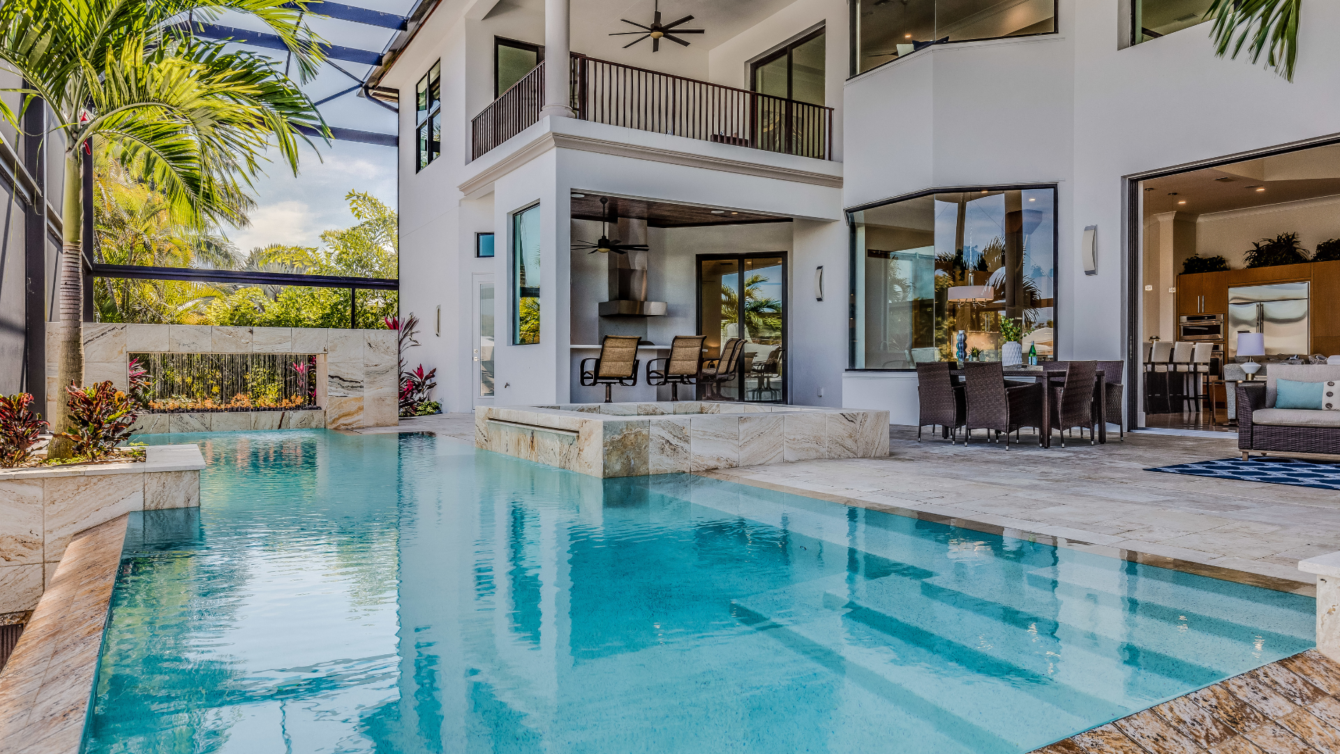 Modern villa with a turquoise backyard pool, stone deck, and open patio seating under tropical trees