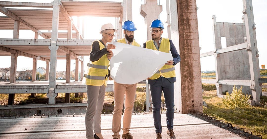 Three construction workers in safety gear reviewing blueprints at a construction site.