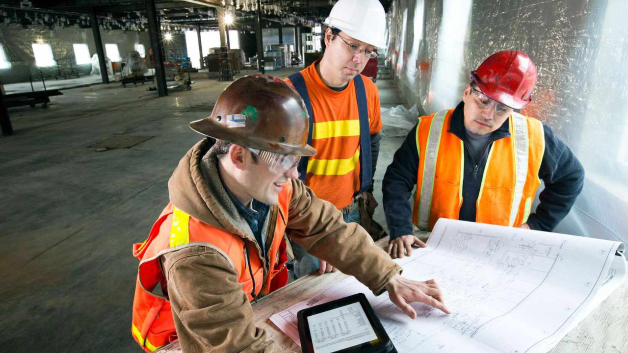 Construction workers in hard hats and vests review blueprints at a job site.