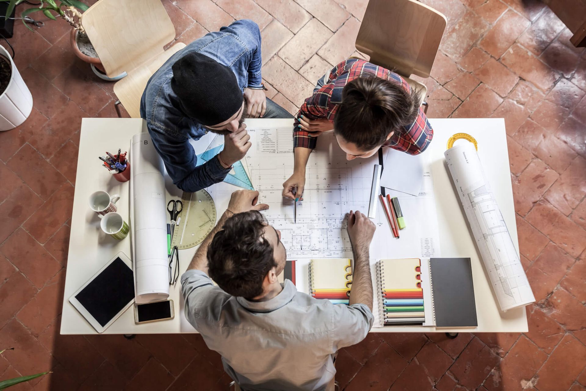 Three people collaborate around a table with blueprints, samples, and tools.
