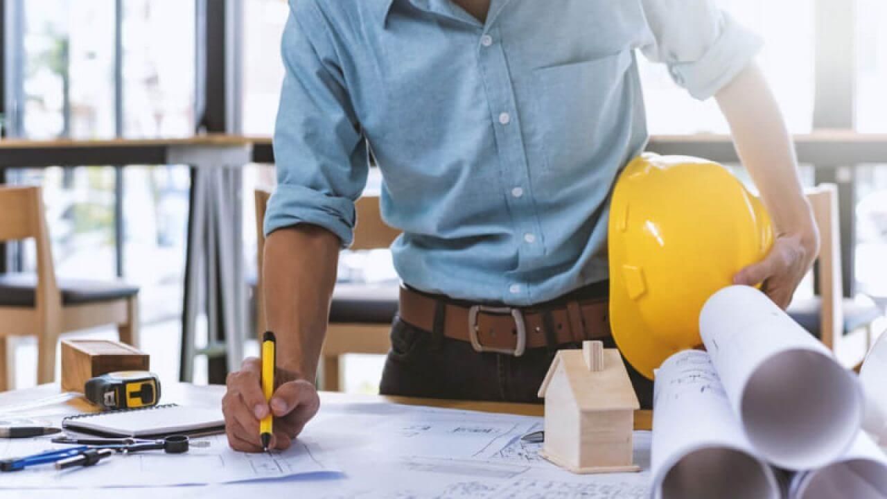 Architect at a table, drafting plans, holding a yellow hard hat, wooden house model, blueprints.