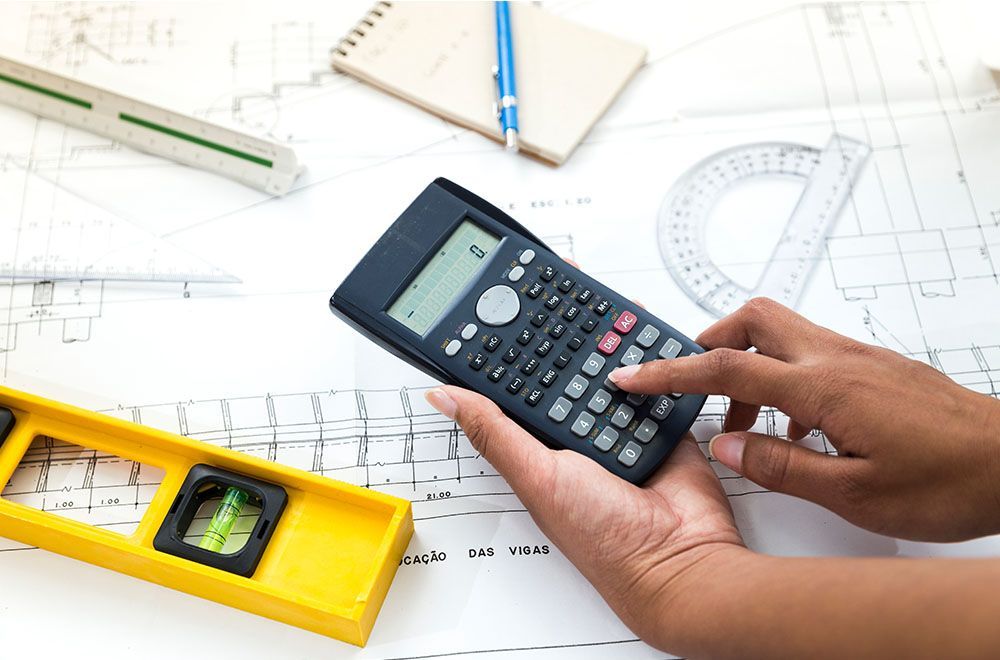 Person using a calculator on a blueprint, with tools like a level and protractor nearby.