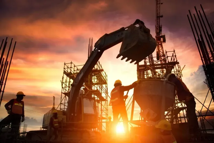 Construction workers on site at sunset; excavator pouring concrete, with orange and yellow hues.