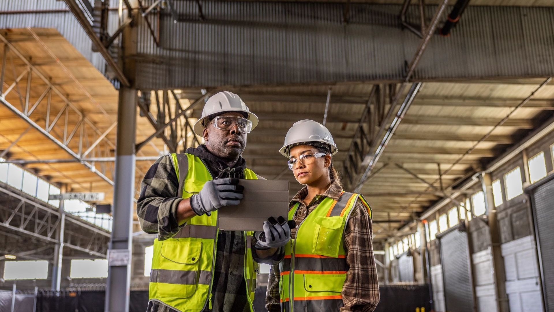 Two construction workers review plans in a warehouse, wearing helmets and vests.