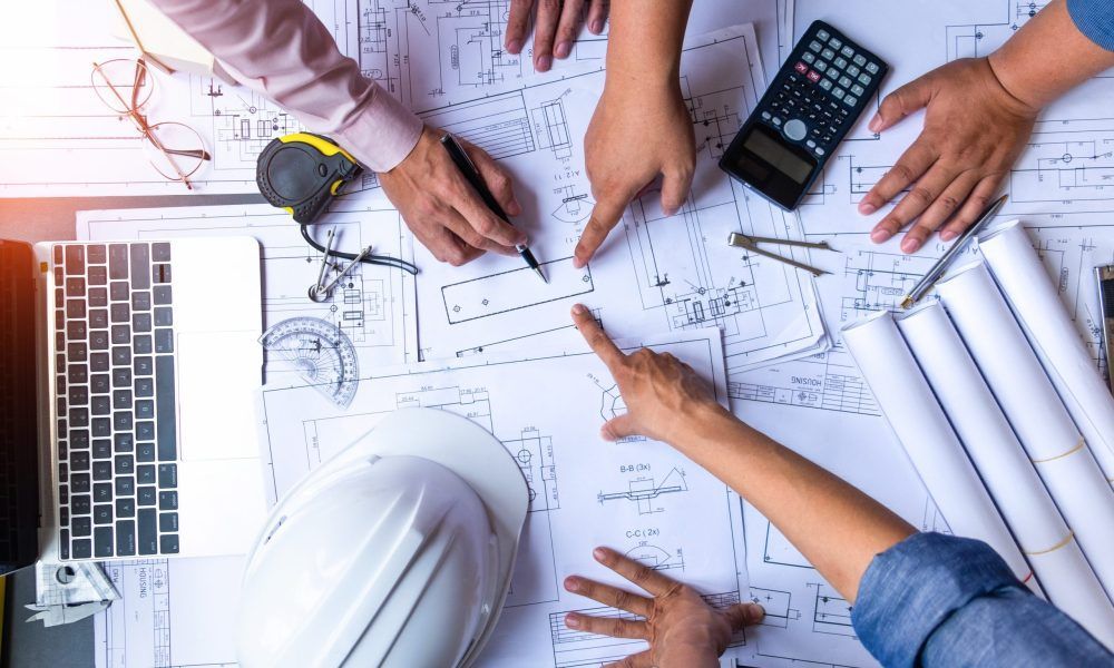 Hands pointing at blueprints on a desk, with a hard hat, laptop, calculator, and rolled plans.