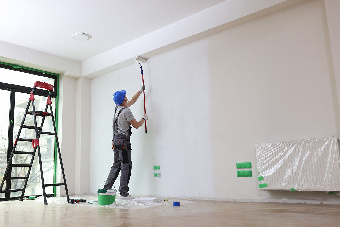 Painter in blue hat using a roller to paint a white wall. Green ladder, open windows, and paint can visible.