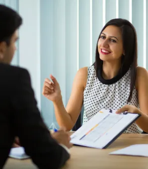 A woman is sitting at a table talking to a man while holding a clipboard.