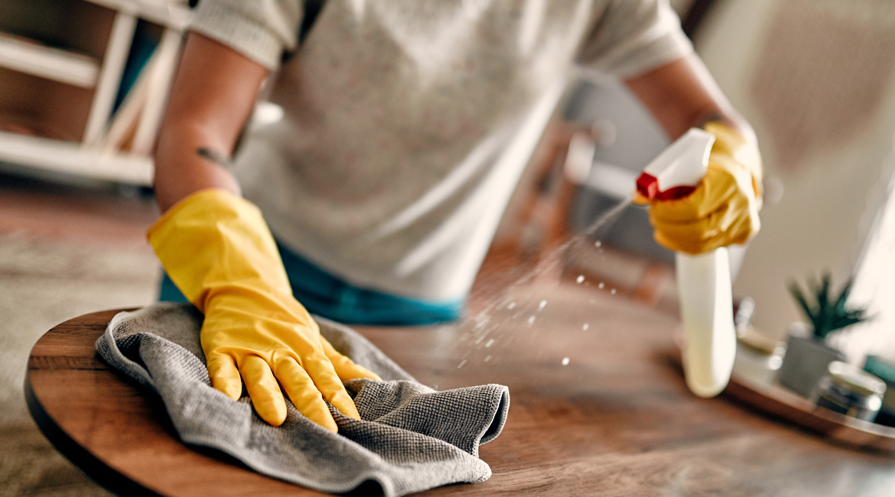 a person wearing yellow gloves is cleaning a wooden table with a cloth and spray bottle .