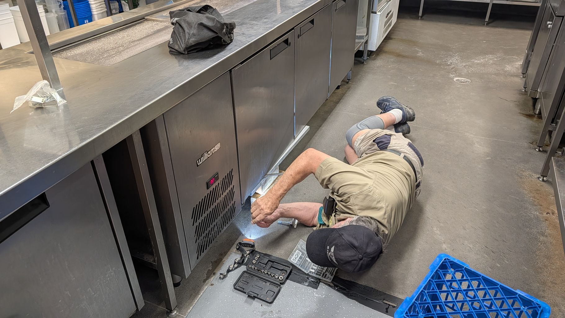 Person Lying on a Kitchen Floor Beside Metal Counters and a Blue Crate — Ace Fridge Door Seals In Cairns, QLD