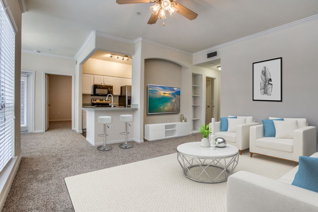 Living room with white furniture, TV, and open kitchen. Brown carpet and neutral walls.