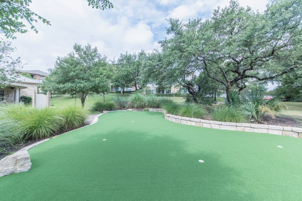 Outdoor community space with a green putting green, stone edging, and trees.