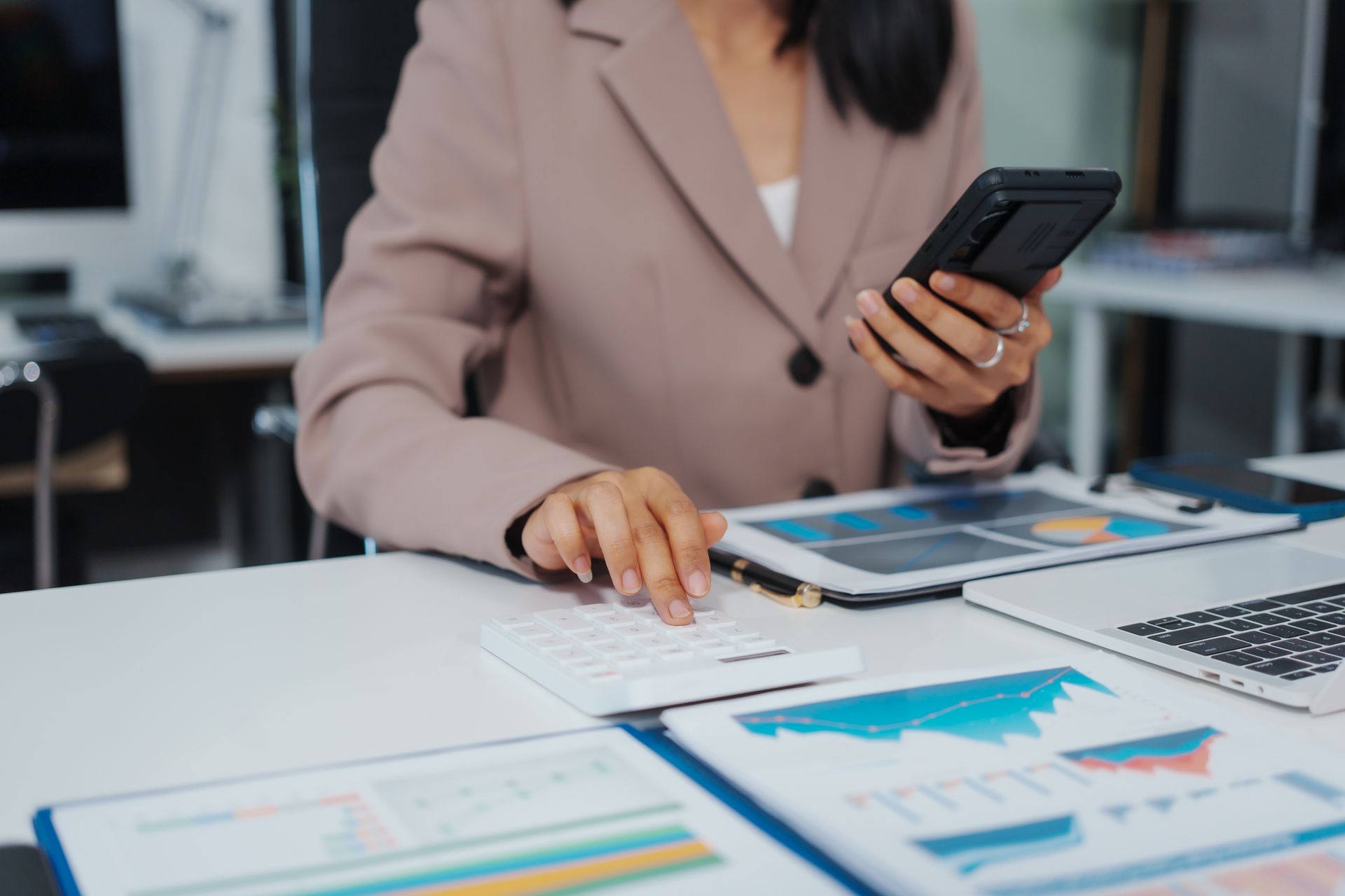 Businesswoman using calculator and phone at desk with charts and laptop.