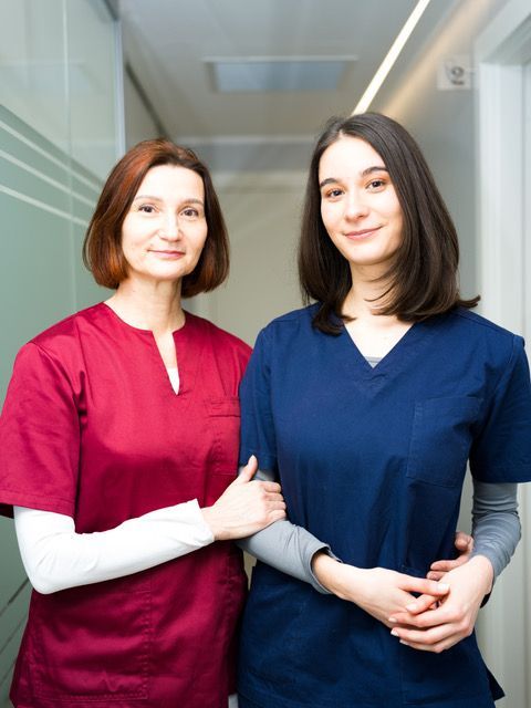 Due professionisti del settore medico, uno in camice bordeaux e l'altro in camice blu scuro, sono in piedi uno accanto all'altro in un corridoio, in posa per un ritratto.