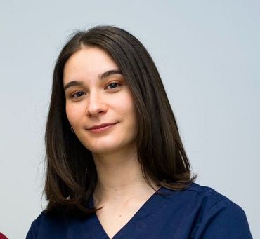 Una persona con i capelli scuri lunghi fino alle spalle indossa una casacca chirurgica blu scuro su uno sfondo grigio chiaro uniforme.