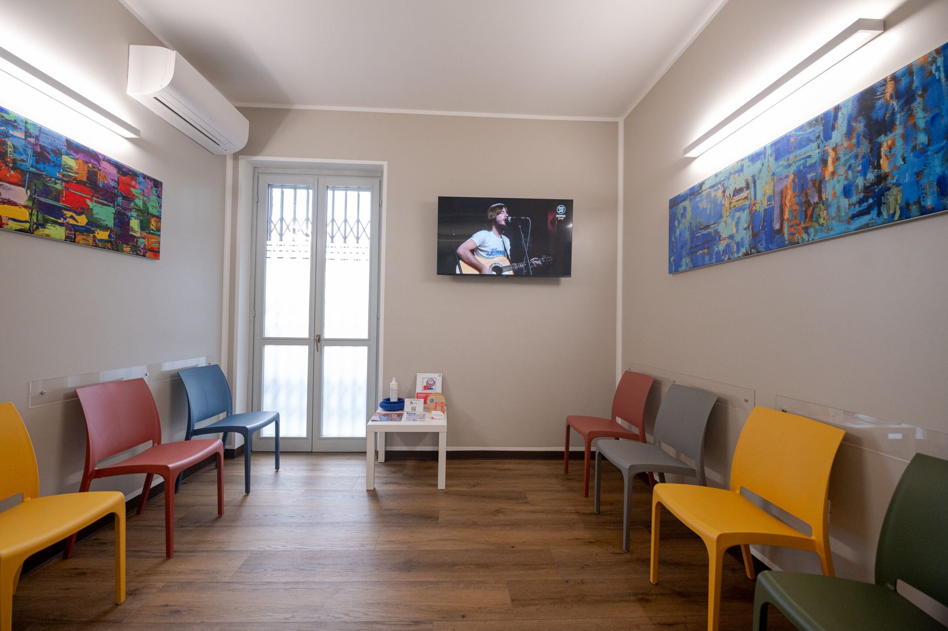 Una sala d'attesa con sedie colorate disposte lungo due pareti, un televisore centrale e opere d'arte alle pareti, il tutto illuminato da una luce calda.