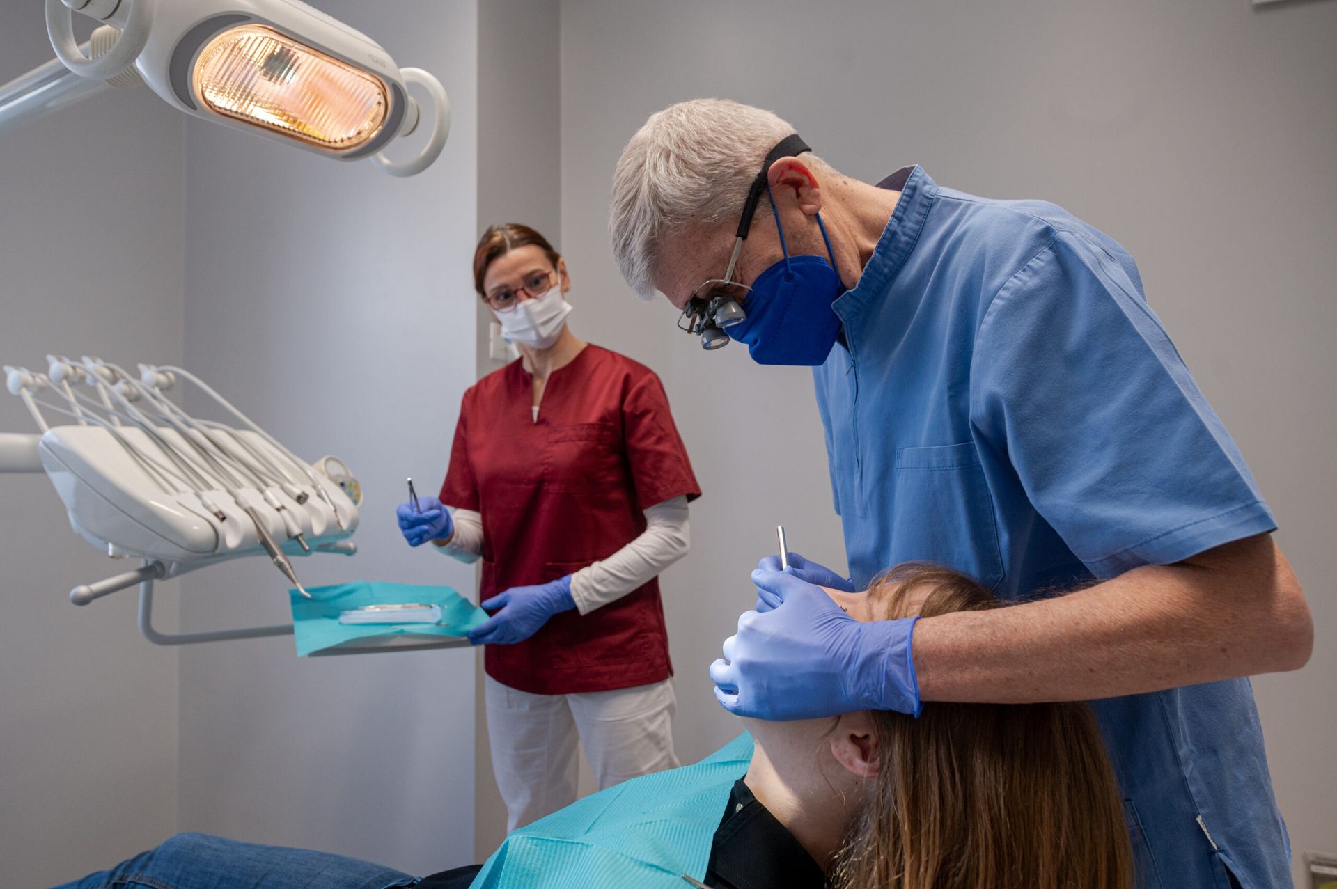 Un dentista, con indosso una divisa blu e una mascherina, lavora su un paziente seduto sulla poltrona odontoiatrica, assistito da un collega con una divisa rossa.