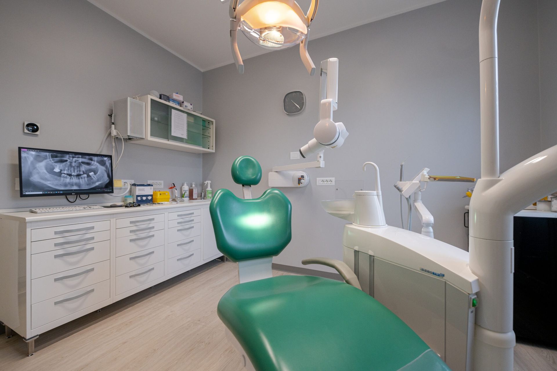 Una luminosa sala per visite odontoiatriche con una poltrona reclinabile verde, attrezzature mediche, uno schermo che mostra una scansione e armadietti bianchi.