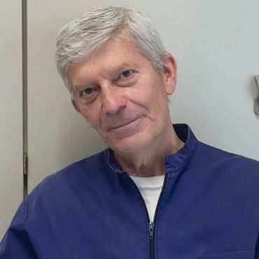 Una persona con i capelli grigi che indossa una giacca blu con cerniera sopra una camicia bianca, guarda direttamente in camera.