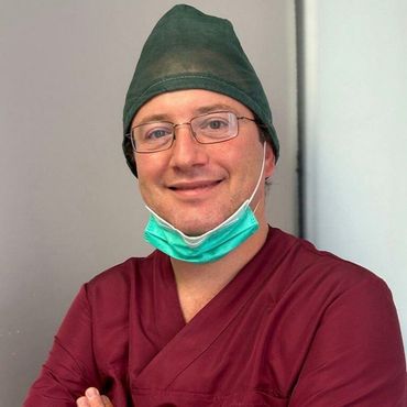 Una persona con indosso una divisa medica rosso scuro e una cuffia chirurgica verde, con la mascherina abbassata, sorride davanti a una parete spoglia.