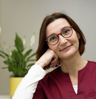 Una persona sorridente con indosso una casacca da infermiere color bordeaux appoggia il mento sulla mano accanto a un vaso di spatifillo.