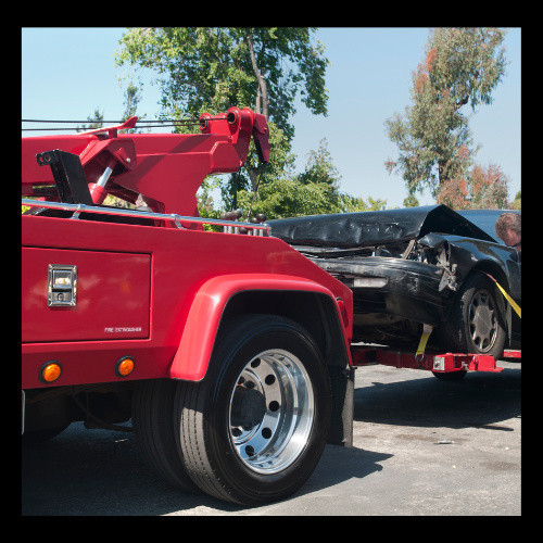 Red tow truck loading a heavily damaged black car. Sunny, outdoors.