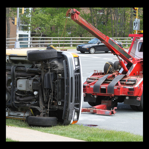 Tow truck lifting a flipped over vehicle at an intersection; red truck, black car, green grass.