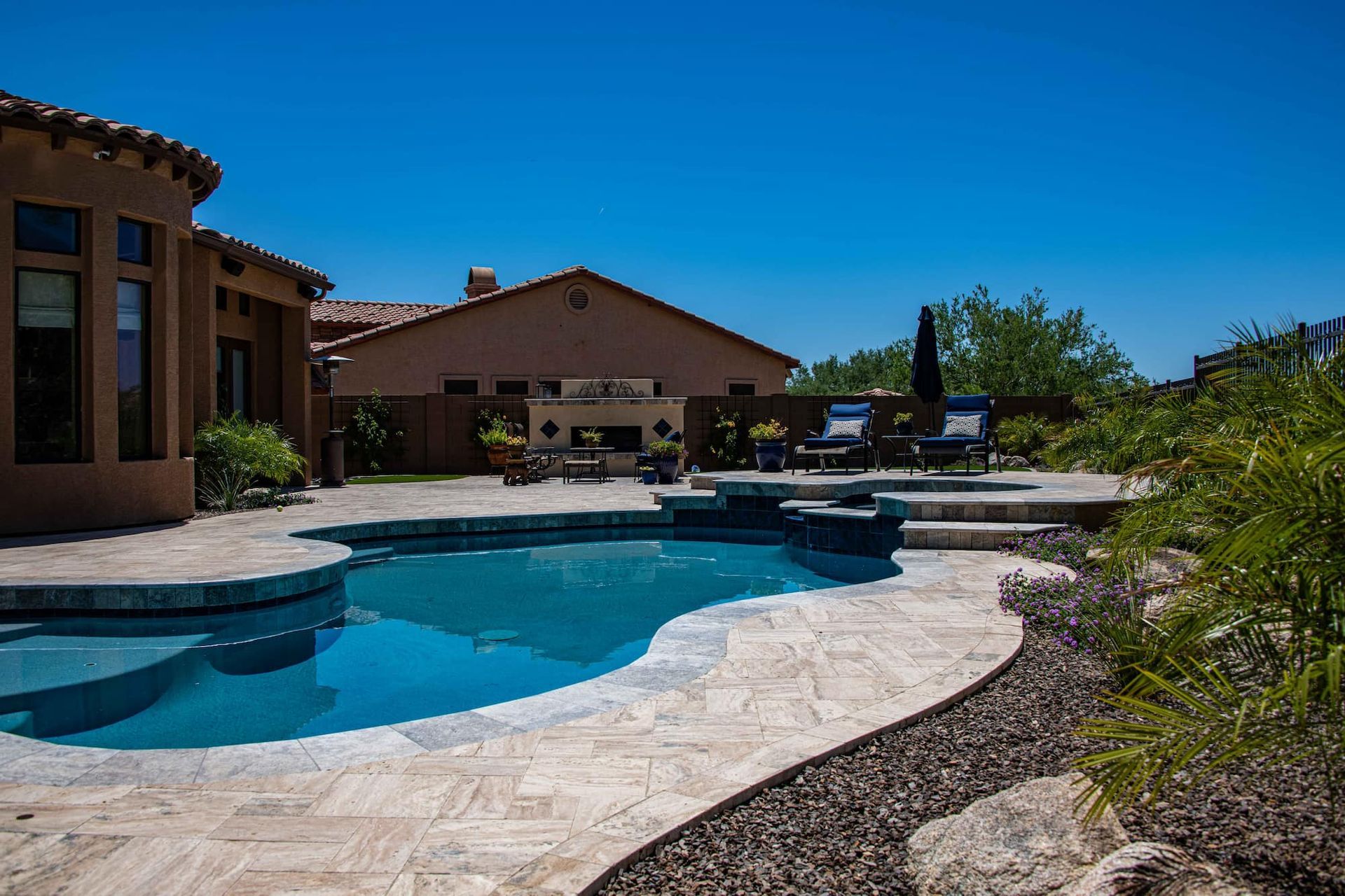 Backyard pool with stone patio, lounge chairs, and house under a bright blue sky.