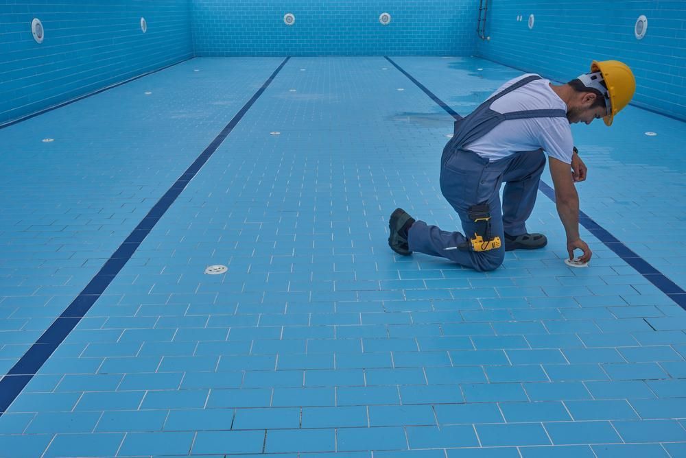 Worker in yellow hard hat kneels in empty blue pool, inspecting the surface.