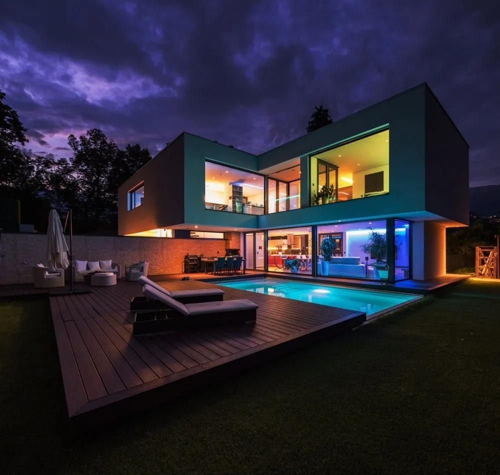 Modern house with pool illuminated at night; deck, lawn, and colorful interior lights.