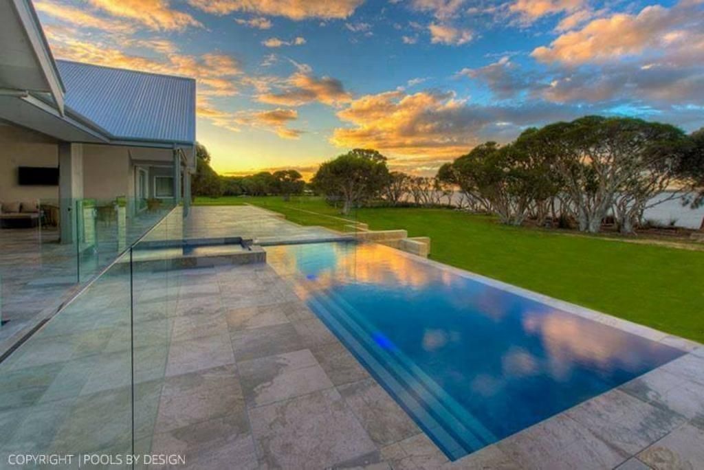 Long rectangular pool reflects a sunset sky; modern house with glass railings on a grassy lawn.