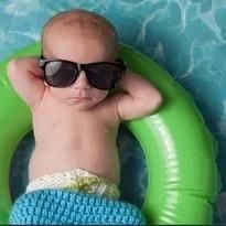 Baby in sunglasses, wearing diaper, lounging on green inflatable ring in water.